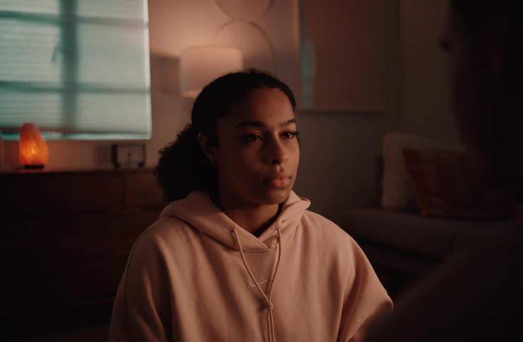 A medium shot focuses on a young woman sitting in a dimly lit room with a soft orange glow from a lamp on a side table a persons hand reaches into the frame to gently touch her chin and she closes her eyes