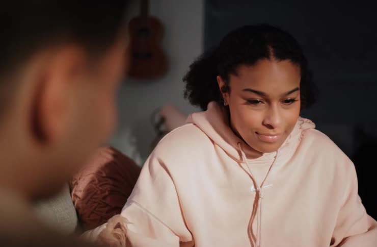 A young woman wearing a pink sweatshirt is engaged in a conversation looking down and then up from an overtheshoulder perspective with a blurred figure in the foreground and an acoustic guitar hanging on the wall behind her