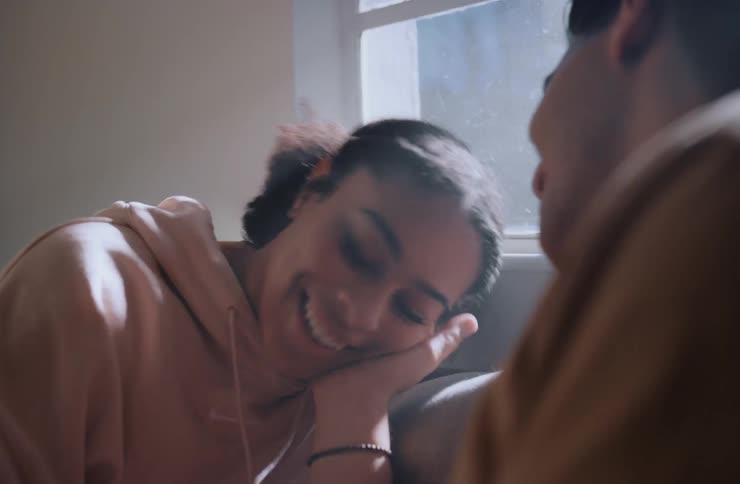 A young man rubs his girlfriends face as they sit on a couch at midday laughing and looking at each other