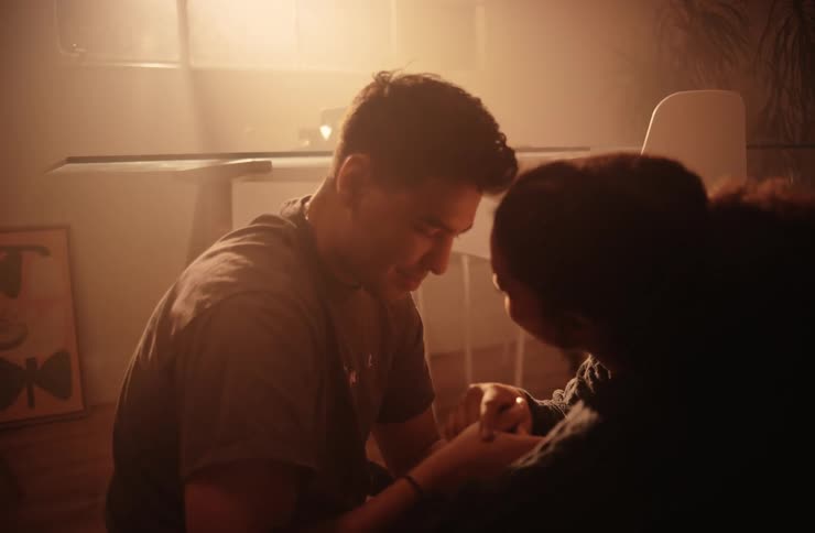 A young adult man in a light colored shirt holds the hands of a young adult woman with dark curly hair sharing a tender moment in a dimly lit room with furniture and light filtering in the background