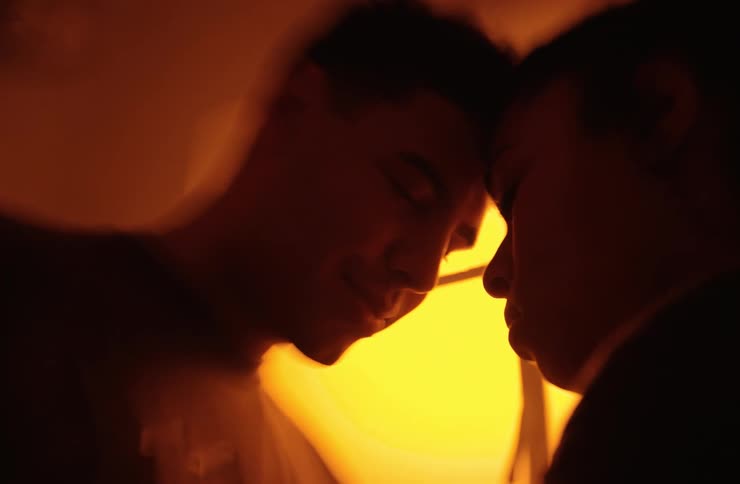A closeup of a couples faces as they stand in a dimly lit room with an orange glow they are touching foreheads and looking at each other sharing an intimate and emotional moment