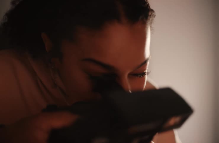 A young woman is looking down with a gentle smile on her face she then lifts a camera to her eye and looks through the viewfinder appearing to be taking a picture