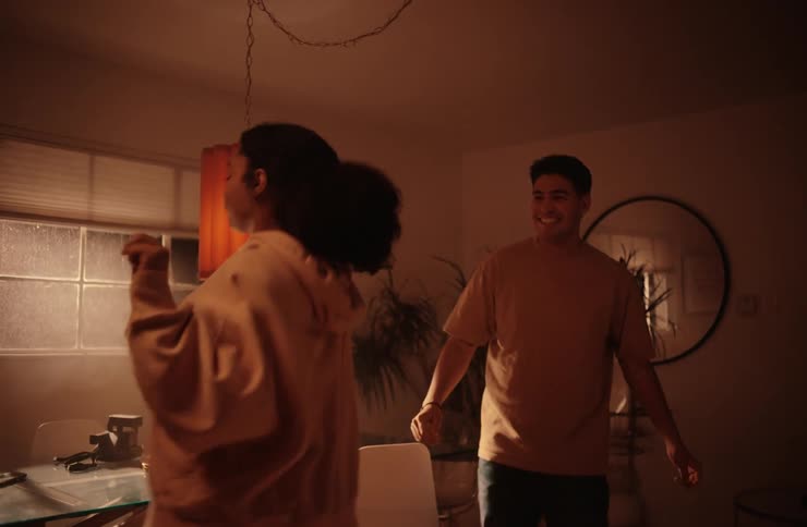 A young happy couple a woman in a light brown hooded sweatshirt and dark pants dances with a man in a brown t shirt and dark pants in a softly lit room