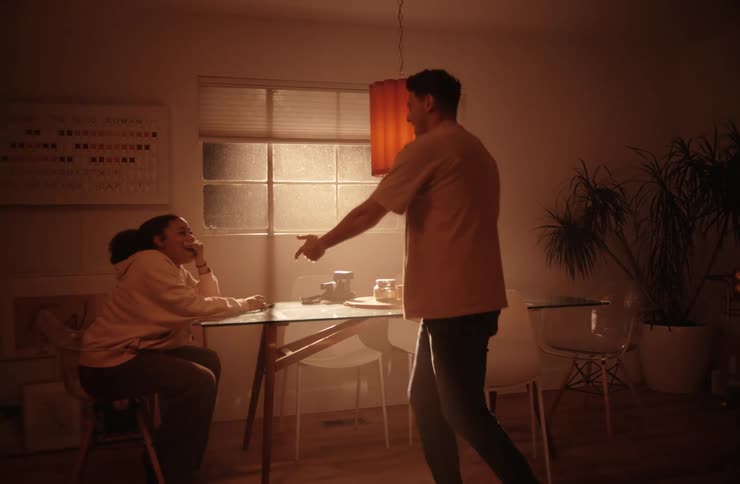 A young man and woman stand and dance together in a dining area indoors