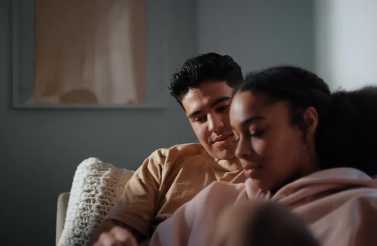 A couple sits closely on a couch with the mans arm around the womans shoulder