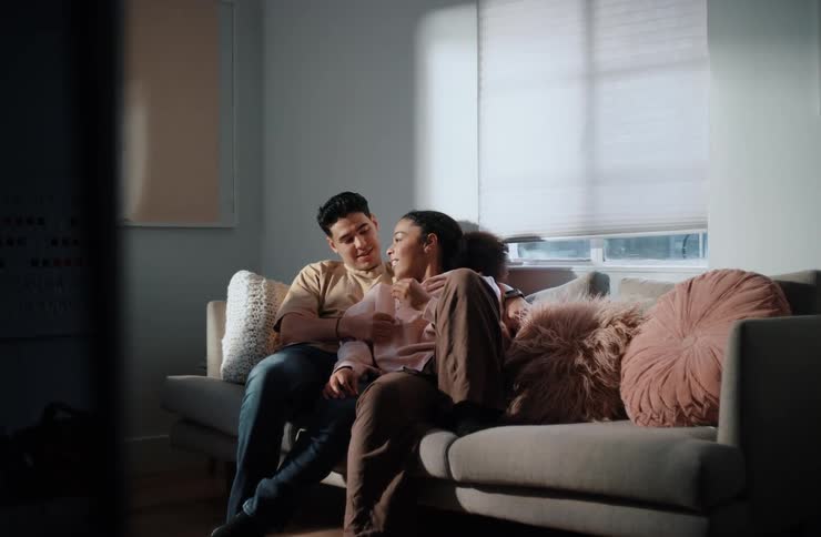 A couple relaxes on a grey sofa lit by natural light from a window to the right the man in a light brown shirt and blue jeans with his arm around the woman in a light pink hoodie and dark pants as they lean into each other
