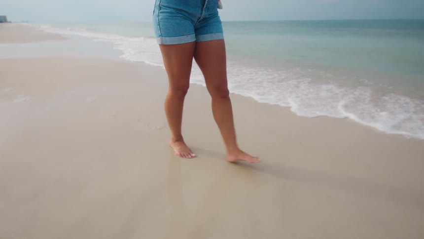 A mediumshot of a woman wearing denim shorts walking barefoot on a wet sandy beach as a gentle wave washes in