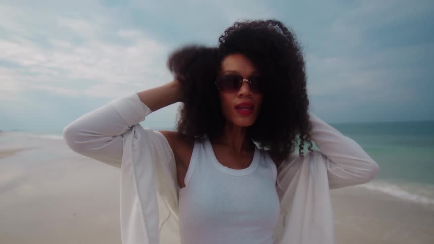 A young woman wearing sunglasses with curly hair and a white shirt poses on the beach sand at midday