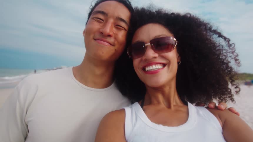 A young woman wearing a white shirt curly hair and sunglasses briefly stood beside a young man as they both smiled at the beach at midday