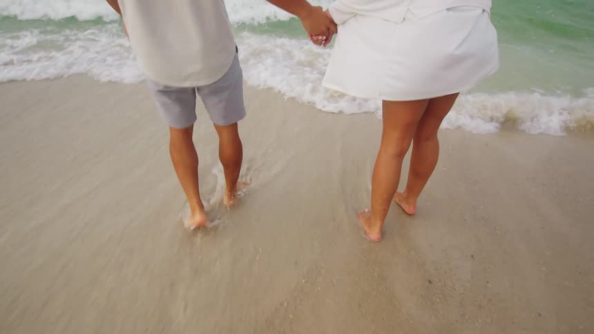 A man and a woman hold hands while walking along a sandy shoreline playing with the waves washing over their bare feet