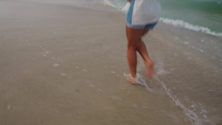 A young woman is happily running barefoot on a sandy beach with her hair flowing and a white shirt trailing behind her as gentle waves lap at her feet