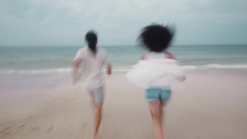 The video shows a blurry shot of two people from behind running towards the ocean on a sandy beach one person is wearing a white shirt and gray shorts while the other has dark curly hair and is wearing a white top with blue shorts