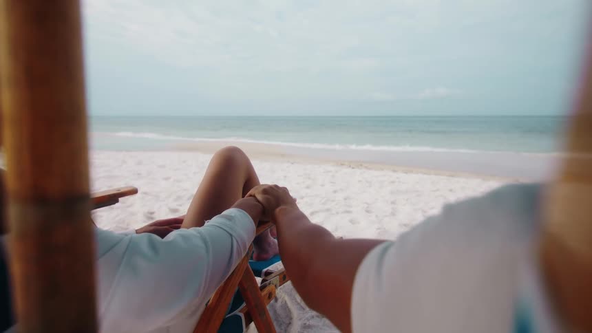 A man and a woman relax on a white sandy beach seated in blue and wooden beach chairs with their hands clasped as a light blue ocean extends to the horizon