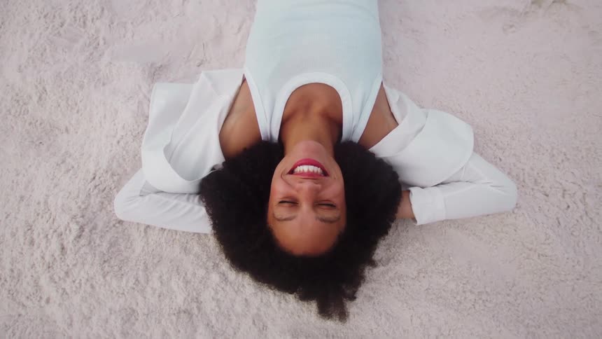 A woman with long dark curly hair smiles and laughs while lying on her back on a white sandy surface with her hands behind her head