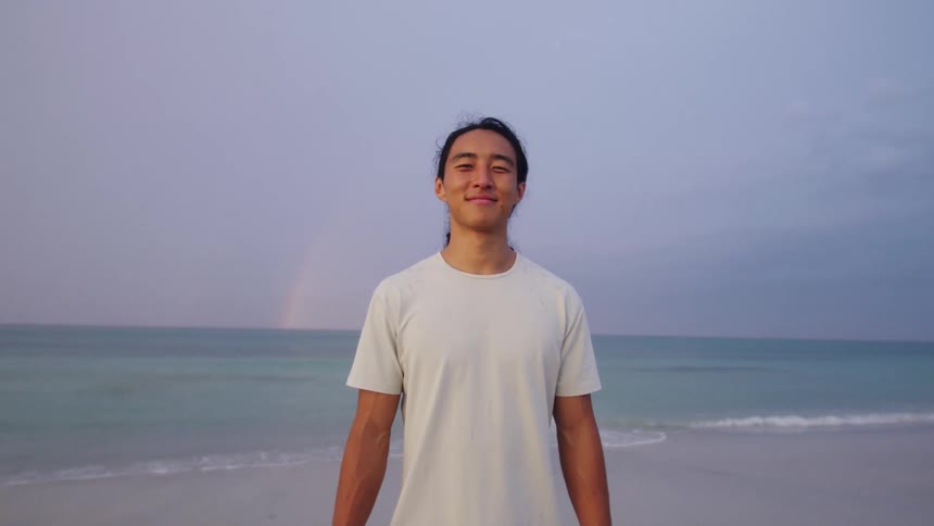 A smiling man with long dark hair stands on a sandy beach wearing a light colored t shirt with a faint rainbow visible over the calm ocean and overcast sky behind him