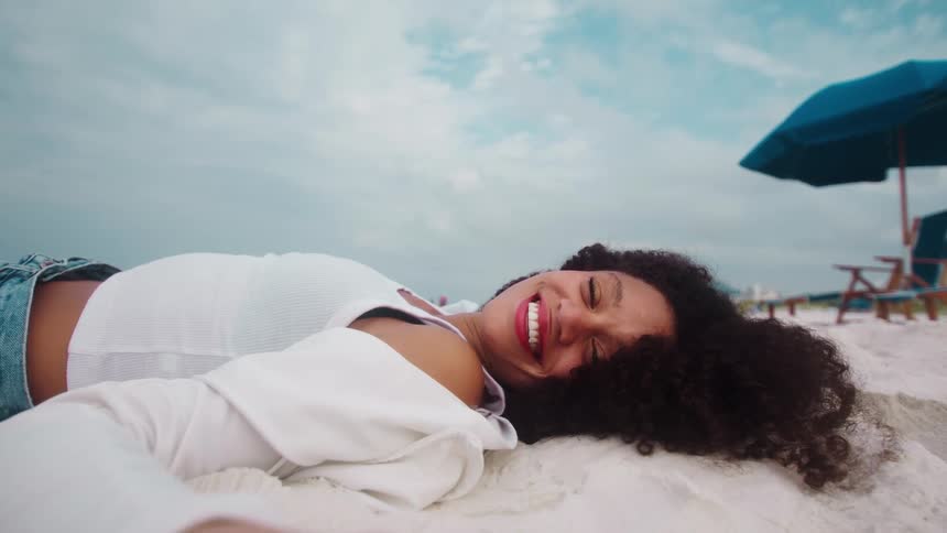 A person is lying on a white sandy beach laughing and smiling widely their hair is a mass of curly black hair spread out on the sand as they film themselves with an outstretched arm