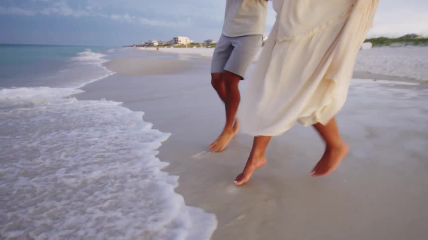 A couple walking barefoot on the beach toward the ocean suddenly jump backward to avoid the waves touching their feet at midday