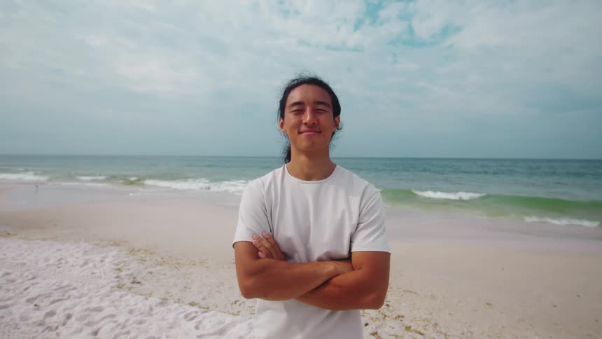 A young man standing on the beach wearing a white tshirt smiling at midday