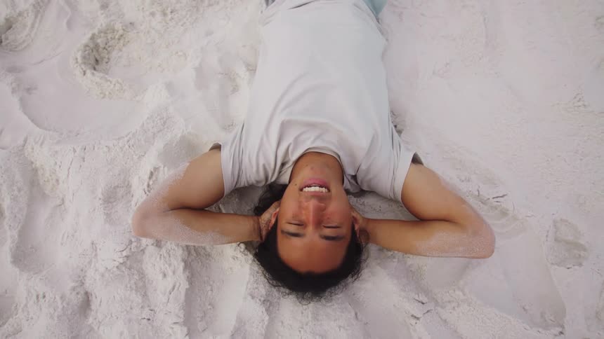 A topdown shot of a man in a white tshirt happily relaxing on his back in the sand with his hands behind his head