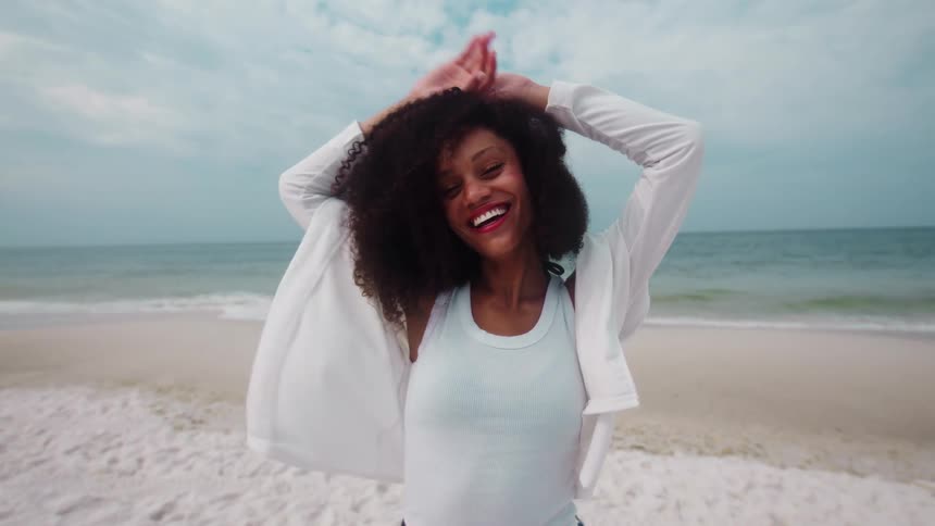A joyful woman with dark curly hair wearing a white tank top and a white long sleeve shirt stands on a sandy beach with gentle waves in the background and raises her hands to her head
