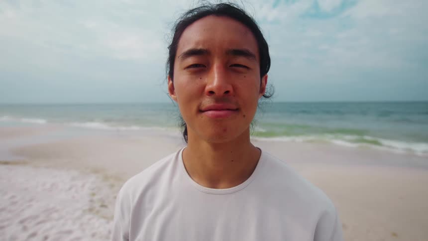 A person with long hair stands on a beach looking directly at the camera with a neutral expression the person smiles and appears to be enjoying the ocean breeze