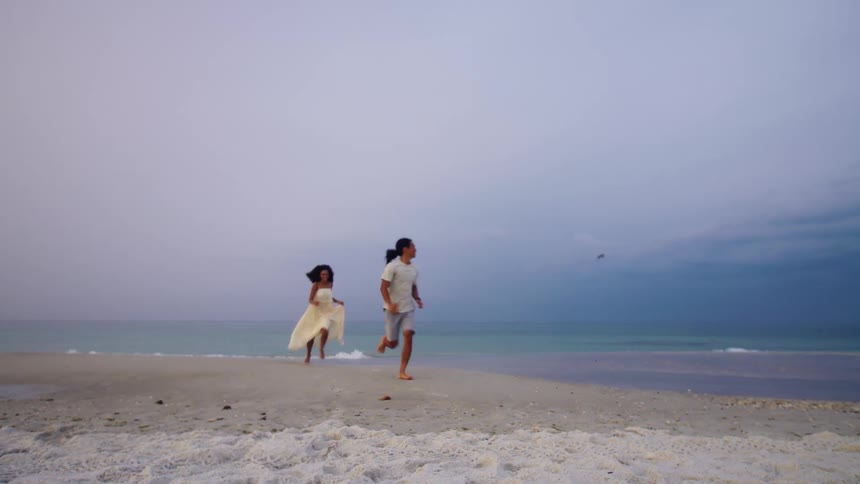 A man in a light t shirt and shorts and a woman in a light colored dress start running toward the camera on a white sandy beach under a cloudy sky