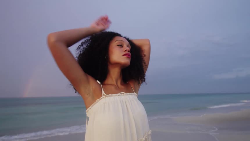 A young woman with curly hair standing and posing on the beach at midday