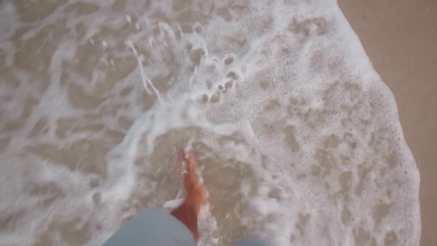 The video shows a firstperson perspective of a person walking on a sandy beach waves can be seen washing over their feet and ankles as they walk