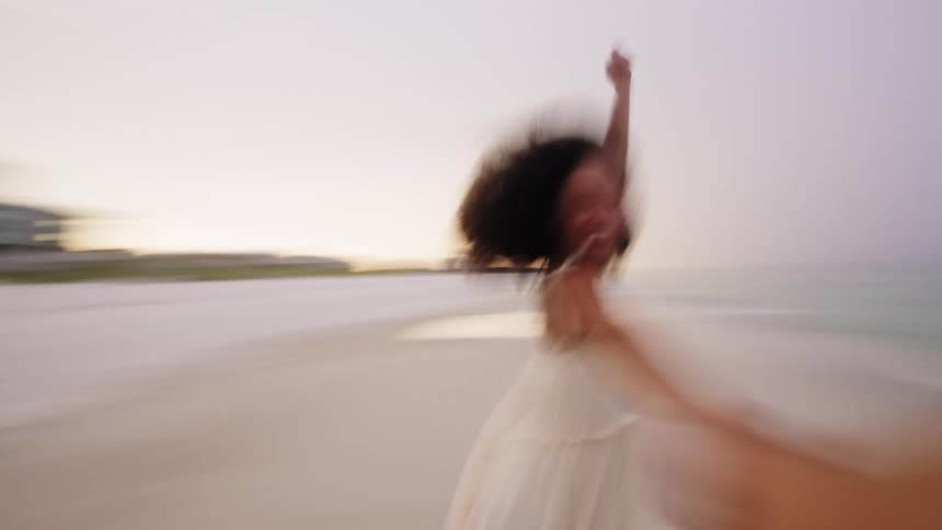 Woman smiling in a white dress spins while holding hands with someone at the beach
