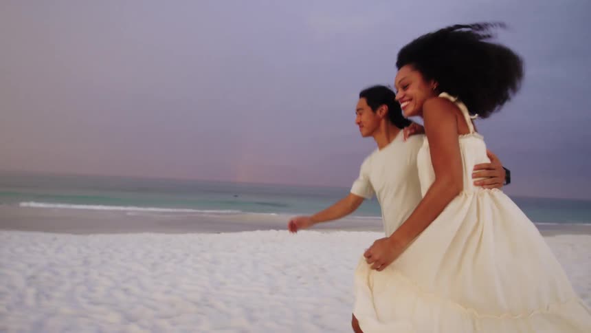 A smiling couple walks handinhand along a beach the woman wearing a lightcolored dress runs ahead playfully with the man following close behind laughing they stop and embrace with their arms around each other