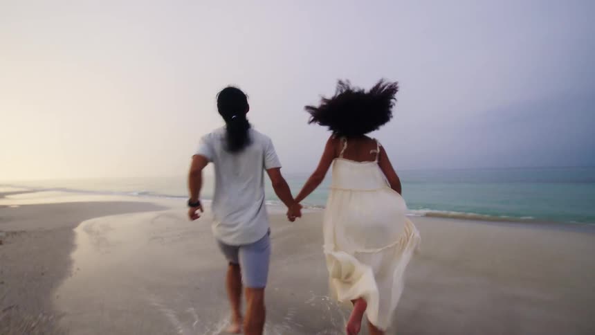 A couple running toward the ocean on the beach holding hands at midday