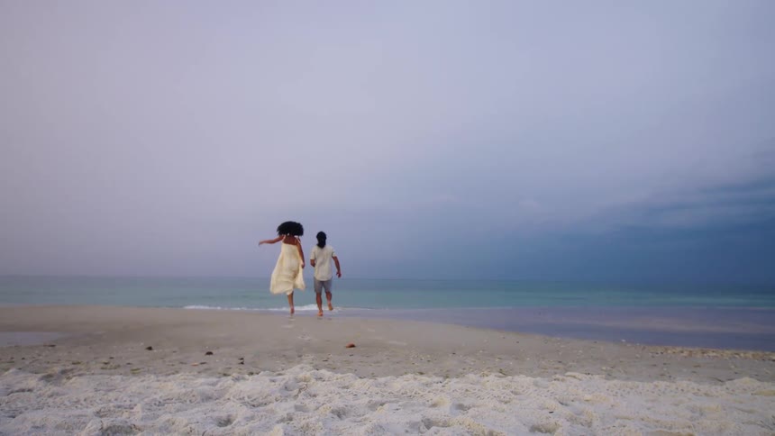A person in a flowing white dress and a man in shorts run towards the water on a beach they stop turn to face the ocean and stand sidebyside enjoying the moment