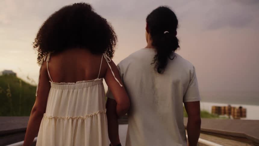 The video shows two people a man and a woman from behind walking on a wooden walkway towards a beach the woman is wearing a white dress and the man is wearing a white tshirt