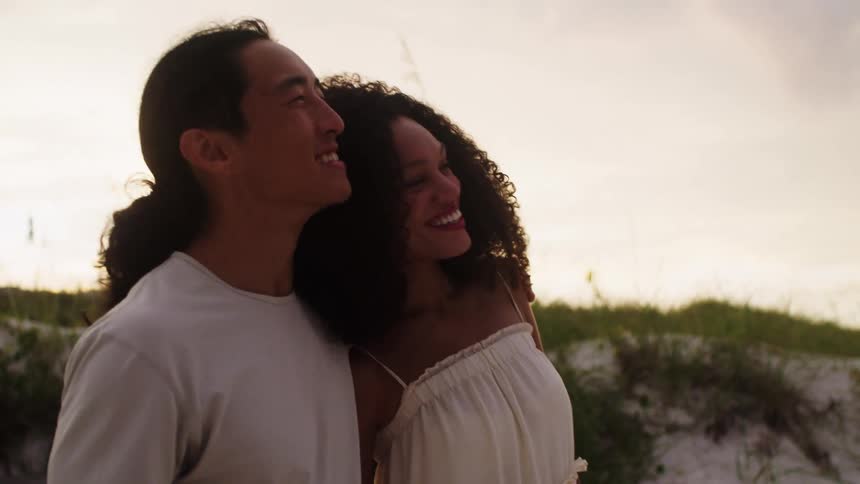 A couple walks along a sandy path beside dunes the man with long dark hair pulled back wearing a light colored shirt placing his arm around the woman with dark curly hair wearing a light colored sundress as they both smile and look forward
