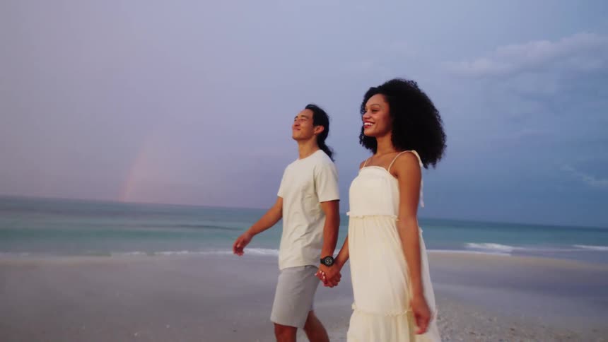 A smiling couple is holding hands while walking along a sandy beach the waves are gently rolling in behind them and the sky is overcast