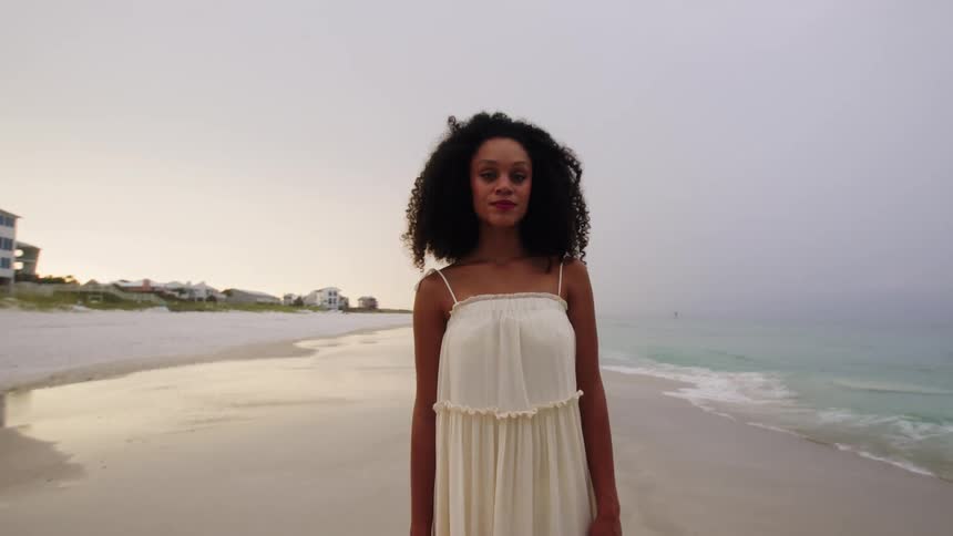 A young woman in a flowing white dress stands on a sandy beach she looks out at the ocean then turns her gaze toward the camera with a neutral expression