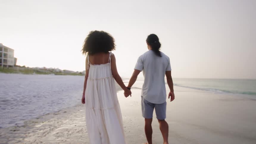 A romantic couple holding hands walks along the beach at sunset