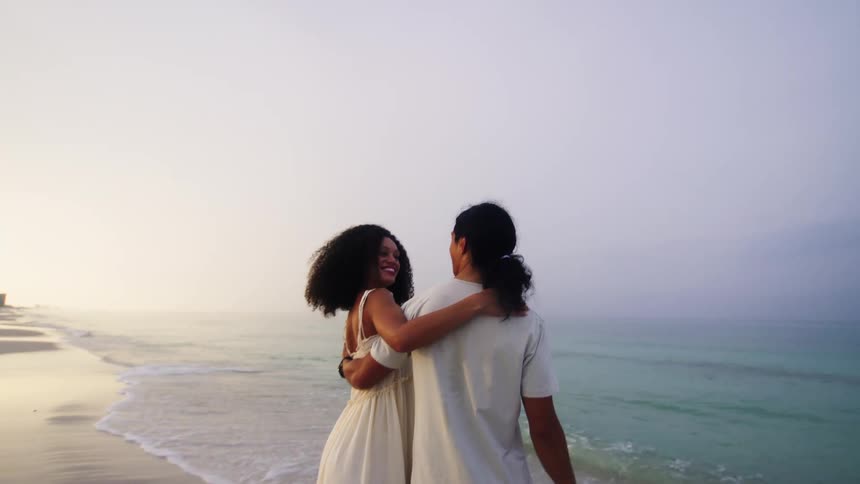 A couple stands on a misty beach with their backs to the camera embracing each other they then turn to look at the camera with the woman smiling as they continue to hold each other