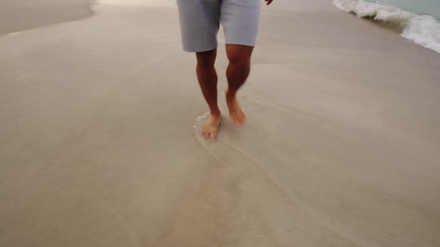 A shot from the knees down of a person walking barefoot on the sand with the waves washing over their feet they are wearing lightcolored shorts