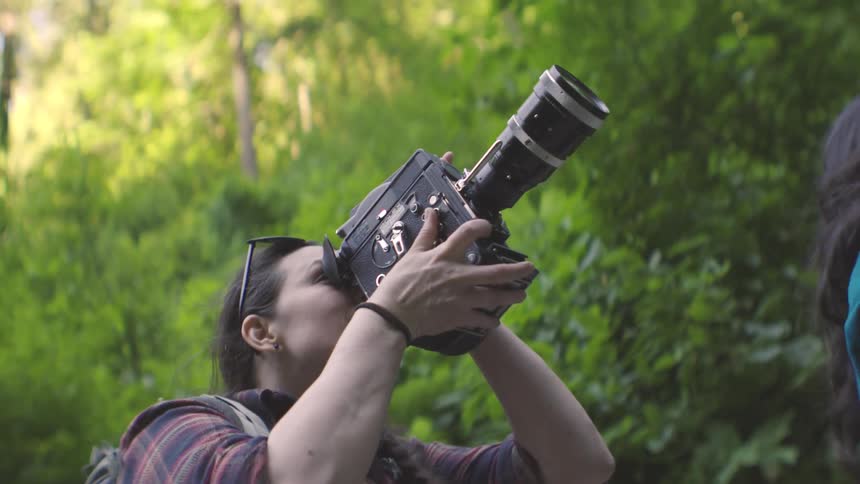 A woman wearing a brown tank top a plaid shirt and a backpack lifts a large black professional camera to her eye to film something upwards in a dense forest