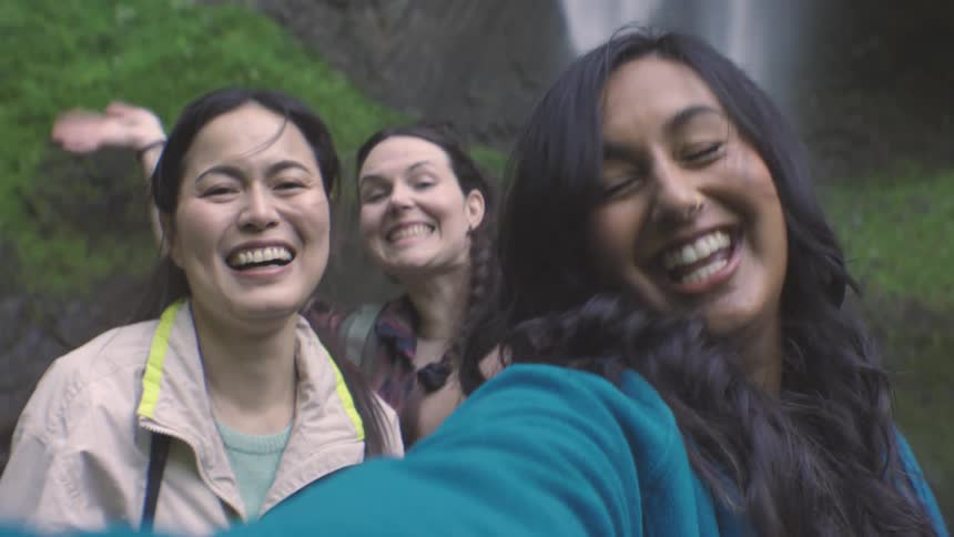 A group of three adult female friends take a selfie outdoors in front of a waterfall during daytime