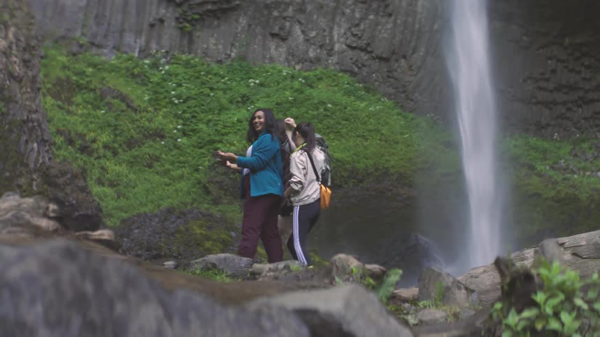 Three women walking on rocks in nature near a waterfall with one of them taking a selfie