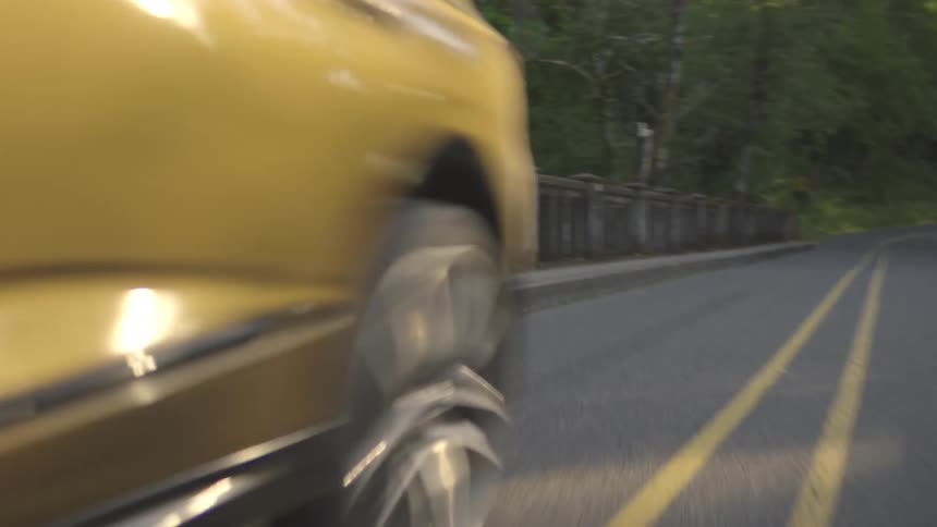 A golden colored suv drives toward the camera on a two lane road with a double yellow line passing by a long ornate stone railing on the left side
