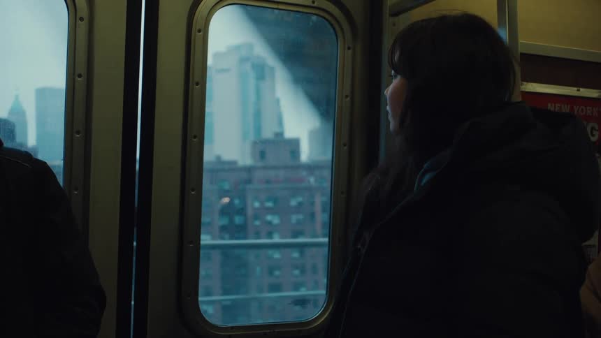 A kid wearing a winter jacket watches through the window of a train as it passes city buildings at dawn