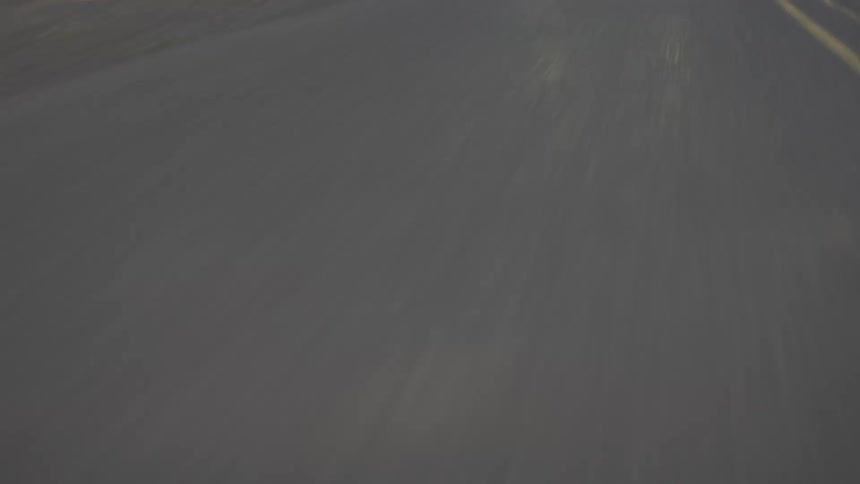 Car is driving ona heavily shaded two lane road the lights are on and the road has lush green bushes and trees on the side of the road