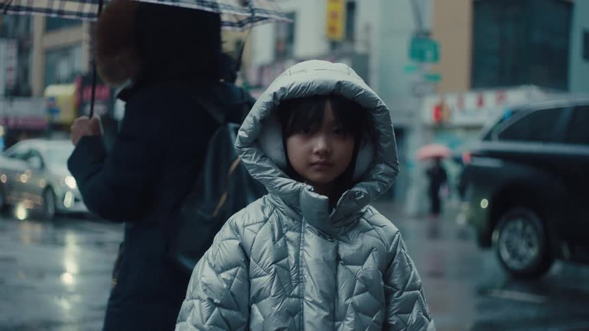 Child wearing a silver puffer jacket with a hood on a rainy city street with cars and pedestrians in the background