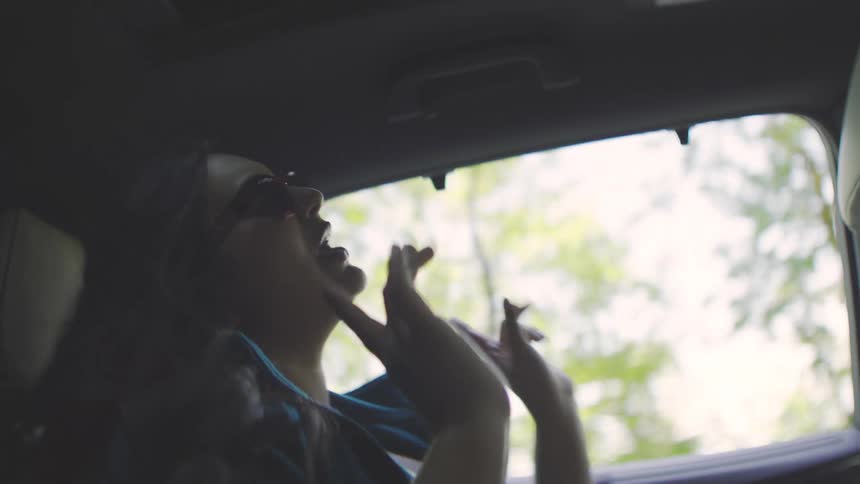 A passenger in a turquoise jacket and sunglasses smiles and dances while singing to the driver of a car as they ride through a forest