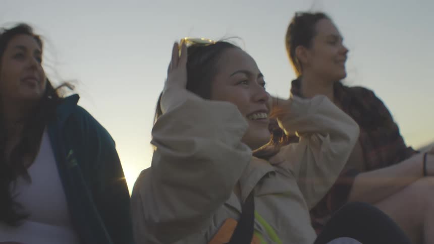 The video shows a group of friends sitting together outdoors as the sun sets one person arrives and joins the group then pulls their hair into a ponytail and smiles