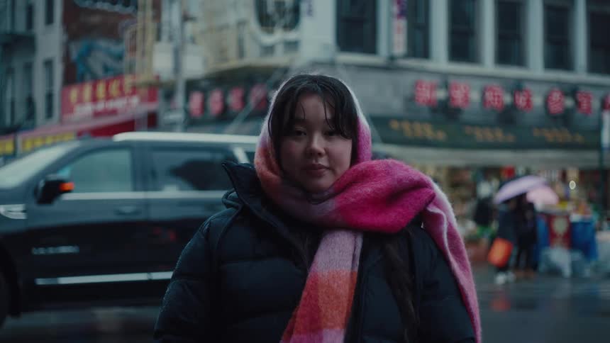 The video shows a medium closeup of a person with dark hair wearing a black puffer jacket and a large pink and orange striped scarf they are standing on a wet city street with cars and other people in the background