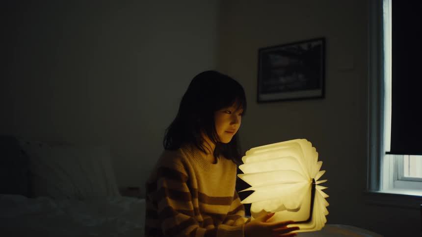 A woman is sitting in a dim room holding an open bookshaped light that illuminates her face she is looking down at the light which is fanned out and glowing brightly